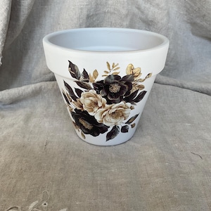 May include: A white tapered plant pot featuring a floral graphic design on the front. The illustration displays a cluster of flowers and leaves in shades of dark brown, cream, and gold, set against the smooth, matte white surface of the container.