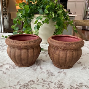 May include: Two round, brown, textured planters with a ribbed body and a decorative embossed rim. The interior of each planter is a smooth, solid red color. These containers feature a wide opening and a tapered base, showcasing a weathered finish.