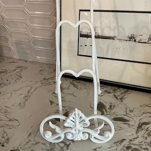 May include: A white metal plate stand featuring a decorative scrollwork base with a central leaf-like motif. The stand has three vertical tiers with heart-shaped tops, designed to hold multiple plates or items in an upright position for display.