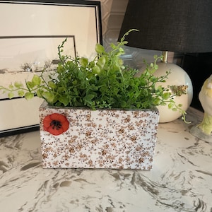 May include: A rectangular white planter box featuring a small, intricate brown floral pattern across its surface and a single, prominent red poppy flower accent on the front left side, filled with lush, vibrant green artificial foliage.