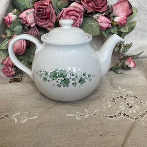 May include: A white ceramic teapot with a rounded body, a curved handle, and a matching lid. The center of the teapot features a decorative horizontal band of green ivy leaves and thin vines painted in a detailed, naturalistic style.
