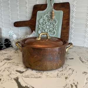 May include: A copper pot with a lid and brass handles sits on a countertop. Behind it are two wooden cutting boards and a decorative, light green floral-patterned cutting board. A white ceramic chicken is also visible.