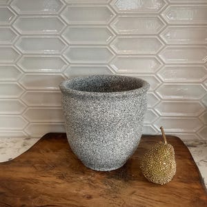 May include: A speckled gray and white cylindrical planter with a rounded rim, accompanied by a small, textured gold pear-shaped decorative object. The planter has a rough, granular surface texture and a wide opening at the top.