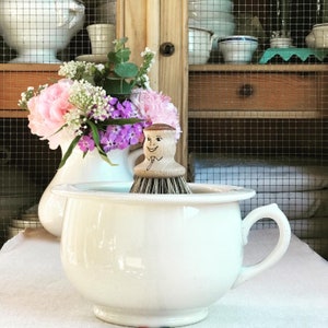 May include: A white ceramic toilet bowl with a wooden brush with a smiling face. The brush is sitting in the bowl. There are pink and purple flowers in a vase behind the bowl.
