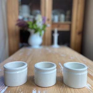 May include: Three small, off-white ceramic jars with a rounded shape and a slightly flared rim are arranged on a light-colored wooden table. The jars are evenly spaced and appear to be empty. The background includes a wooden cabinet and a vase of flowers.