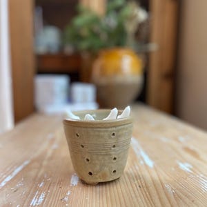 Op de afbeelding: Een kleine, beige keramische pot met een reeks gelijkmatig verdeelde gaten en horizontale groeven. De pot is gevuld met witte voorwerpen. De pot staat op een lichtgekleurd houten oppervlak. Een vage achtergrond omvat een houten kast en een gele en beige pot.
