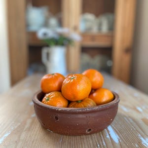 May include: A round, brown, unglazed clay bowl with small circular perforations along the side, filled with several bright orange tangerines. The bowl has a rustic, textured surface and a wide, open top, containing the fresh citrus fruit.