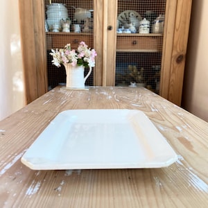 May include: A square, off-white ceramic plate with slightly rounded corners sits on a rustic wooden table. The plate's surface reflects light, suggesting a glossy finish. The background includes a wooden cabinet with glass doors and decorative items.