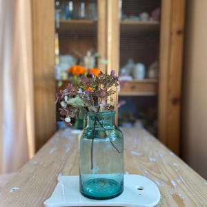 Pode incluir: Um vaso de vidro transparente, cor turquesa, com flores secas, colocado sobre uma bandeja de cerâmica branca. O vaso tem formato cilíndrico com base larga. O arranjo está sobre uma mesa de madeira clara.