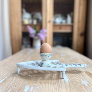 May include: A brown egg in a small, ornate egg cup with a blue and white floral design, set on a white, cast-iron egg rack. The rack has a decorative, openwork design and is placed on a light-colored wooden table.