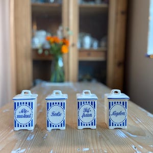 Puede incluir: Cuatro botes de especias de cerámica blanca con rayas verticales azules y texto. Cada bote tiene un asa pequeña en la tapa. El texto en los botes incluye "Note-muskaat", "Foelie", "Stuf-fraan" y "Nagelen".