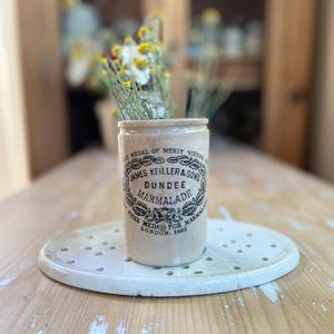 May include: A vintage Dundee Marmalade jar with a floral arrangement. The beige ceramic jar features black text and a decorative emblem. It sits on a white, oval-shaped plate with small holes. The jar contains yellow and white flowers.