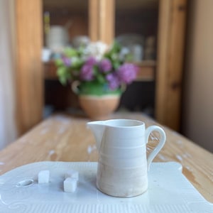 May include: A small, white ceramic creamer with a handle and spout, on a white rectangular tray. The creamer has horizontal ridges and a light brown gradient at the base. Three sugar cubes are on the tray.