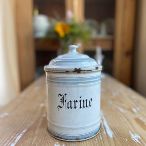 May include: A cylindrical white enamel canister with a matching lid, featuring light blue horizontal stripes and the word Farine printed in black gothic-style lettering. The container shows signs of wear with visible rust along the edges.