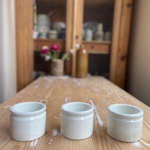 May include: Three small, off-white ceramic jars with rounded bodies and a slightly flared rim sit on a weathered wooden table. The jars are empty and appear to be vintage or antique, with a simple, classic design. The background includes a wooden cabinet.