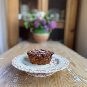 May include: A baked pastry on a white plate with a red floral design. The pastry is golden brown and dusted with powdered sugar. The plate sits on a wooden table. A vase of flowers is in the background.
