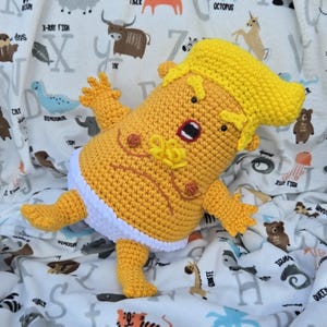 Puede incluir: Muñeca de ganchillo hecha a mano con forma de bebé, con pelo y piel amarillos, y un pañal blanco. La muñeca tiene la boca abierta y está sobre una manta blanca con ilustraciones de animales y el texto "X-RAY FISH".