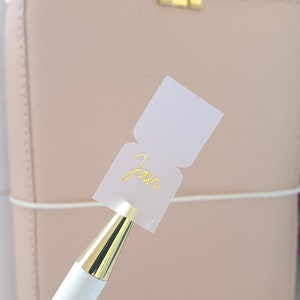 Peut inclure: Un stylo blanc avec un capuchon doré tient un autocollant blanc sur lequel est inscrit le mot "Jan" en lettres dorées. L'autocollant est fixé à un carnet relié en cuir rose.