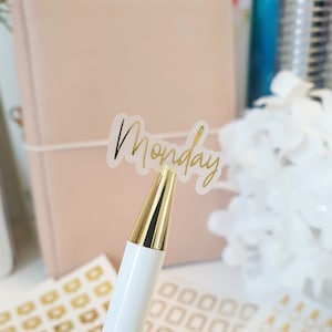 Könnte beinhalten: Ein weißer Stift mit goldfarbener Spitze hält einen transparenten Aufkleber mit dem Wort "Monday" in goldener Schrift. Der Hintergrund umfasst einen rosa Planer und andere Schreibwaren, was auf ein Thema von Organisation und Planung hindeutet.