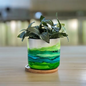 May include: A white ceramic planter with a green, blue, and yellow abstract design. The planter is filled with a green leafy plant and is sitting on a wooden coaster.