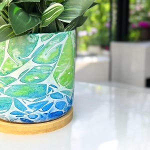 May include: A ceramic planter with a blue, green, and white leaf pattern. The planter is sitting on a wooden coaster.