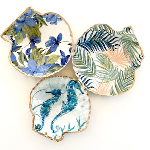 May include: Three decorative ceramic seashell dishes with gold trim. The first dish features a blue floral pattern, the second has a tropical leaf design, and the third has a blue seahorse illustration.
