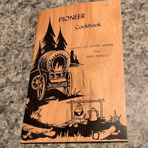 May include: A light brown cookbook with a wood grain pattern featuring black illustrations of a covered wagon, pine trees, and a campfire with cooking pots. The text reads PIONEER Cookbook, CAMPFIRE AND KITCHEN RECIPES From EARLY AMERICA.