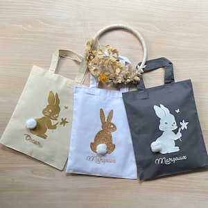 Peut inclure: Trois sacs cabas avec des motifs de lapins. Les sacs sont blancs, beiges et gris. Les lapins sont en paillettes dorées, blancs et en paillettes dorées. Les sacs sont personnalisés avec les noms "Oscar", "Margaux" et "Margaux".