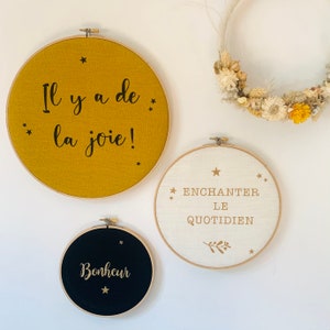 Peut inclure: Trois cerceaux de broderie ronds avec des tissus de couleurs différentes et des lettres dorées. Le cerceau du haut est jaune moutarde avec des lettres noires qui disent "Il y a de la joie !". Le cerceau du milieu est blanc avec des lettres dorées qui disent "Enchanter le quotidien". Le cerceau du bas est noir avec des lettres dorées qui disent "Bonheur".