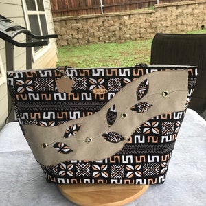 Puede incluir: Una bolsa de lona negra y blanca con un patrón geométrico y una superposición de tela de color canela con recortes de hojas. La bolsa tiene una etiqueta de cuero marrón con la palabra "Onyx" en ella.