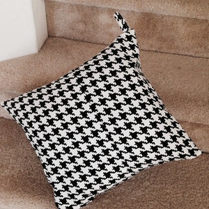 May include: A black and white houndstooth patterned pillow with a loop handle. The pillow is lying on a set of stairs with a brown carpet.