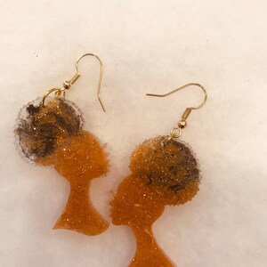 May include: A pair of gold earrings featuring a silhouette of a woman with an afro. The earrings are made of resin and have a glittery finish.