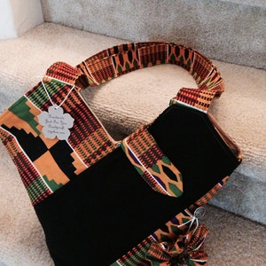 Puede incluir: Una bolsa de tela negra y amarilla con una correa larga. La bolsa tiene un bolsillo en la parte delantera y está decorada con un estampado africano colorido. La bolsa tiene una etiqueta que dice "Handmade Just For You BoutiqueCraft" y una dirección de sitio web.