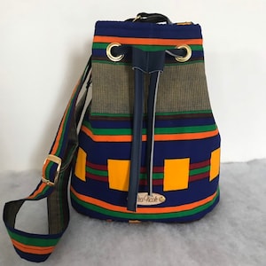 Könnte beinhalten: Eine blaue und orangefarbene Eimer-Tasche mit einem langen Riemen. Die Tasche hat einen Kordelverschluss und goldene Ösen. Die Tasche ist aus Stoff mit einem geometrischen Muster und hat ein Leder-Etikett mit der Aufschrift "La-Nicole".