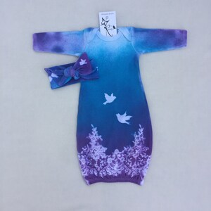 May include: A purple and blue tie-dye baby gown with white birds and floral designs. The gown has long sleeves and a bow headband.
