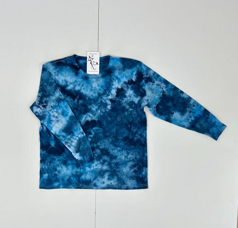 May include: A long-sleeve tie-dye shirt in shades of blue and white. The shirt has a crew neck and a tag attached near the neckline. The tie-dye pattern creates a marbled effect across the fabric. The shirt is laid flat on a white surface.