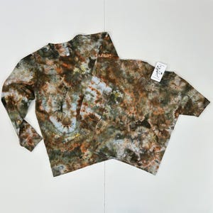 May include: Two tie-dye shirts, one long-sleeved and one short-sleeved, in shades of brown, green, and blue. The shirts have a crew neck and a tag is attached to the short-sleeved shirt. The tie-dye pattern is unique on each shirt.