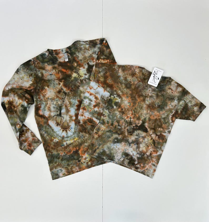 May include: Two tie-dye shirts, one long-sleeved and one short-sleeved, in shades of brown, green, and blue. The shirts have a crew neck and a tag is attached to the short-sleeved shirt. The tie-dye pattern is unique on each shirt.