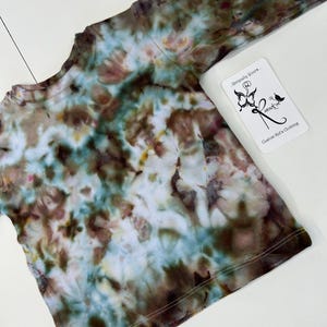 May include: A long-sleeved tie-dye shirt in shades of brown, blue, and white. The shirt has a crew neck and a tag that reads "Uniquely Yours. Custom Kid's Clothing."