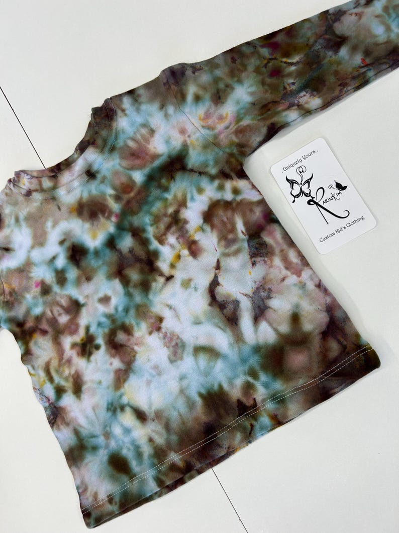 May include: A long-sleeved tie-dye shirt in shades of brown, blue, and white. The shirt has a crew neck and a tag that reads "Uniquely Yours. Custom Kid's Clothing."