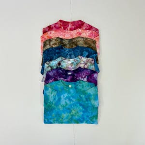 May include: A stack of six tie-dye t-shirts in various colors, including pink, orange, brown, blue, purple, and teal. The shirts are neatly folded and stacked on a white surface. Each shirt has a crew neck and short sleeves.