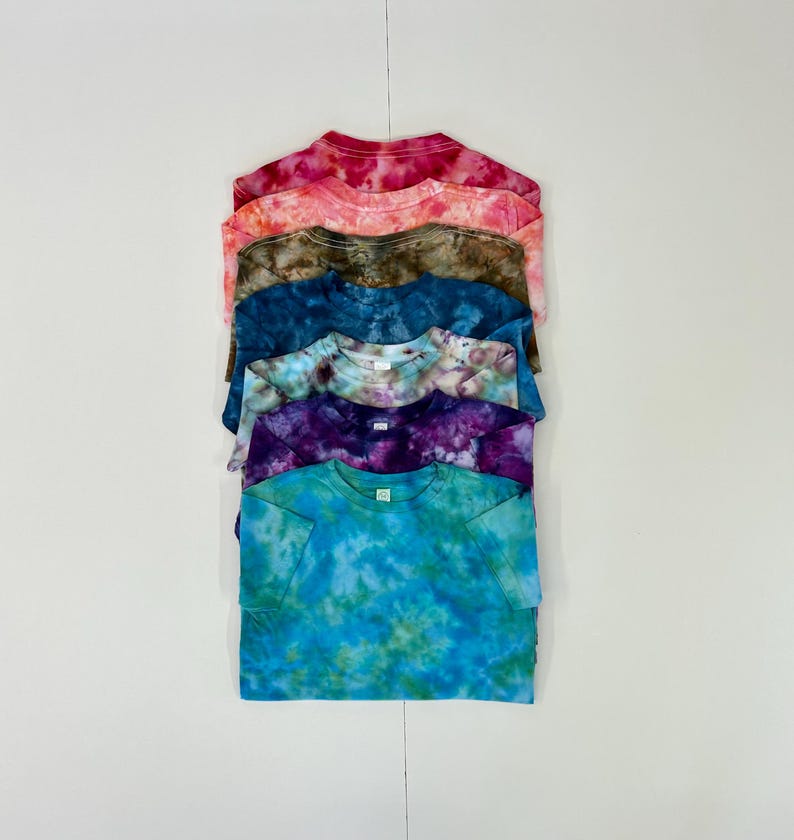 May include: A stack of six tie-dye t-shirts in various colors, including pink, orange, brown, blue, purple, and teal. The shirts are neatly folded and stacked on a white surface. Each shirt has a crew neck and short sleeves.