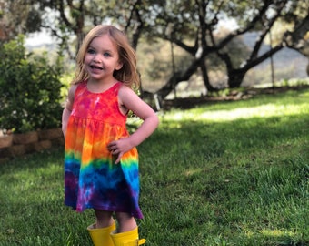 May include: A young girl wearing a rainbow tie-dye dress and yellow rain boots stands in a grassy field.