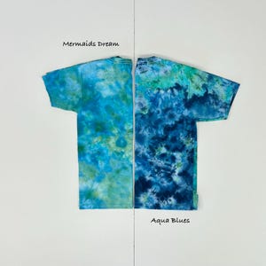 May include: Two tie-dye t-shirts, side-by-side, each with a unique blue and green pattern. The left shirt is labeled "Mermaids Dream," and the right shirt is labeled "Aqua Blues." Both shirts have short sleeves and a crew neck.