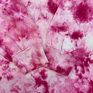 May include: Close-up of a long-sleeved shirt with a tie-dye pattern in shades of pink and white. The cuffs of the sleeves are visible, showing a ribbed texture. The fabric has a soft, blurred appearance.