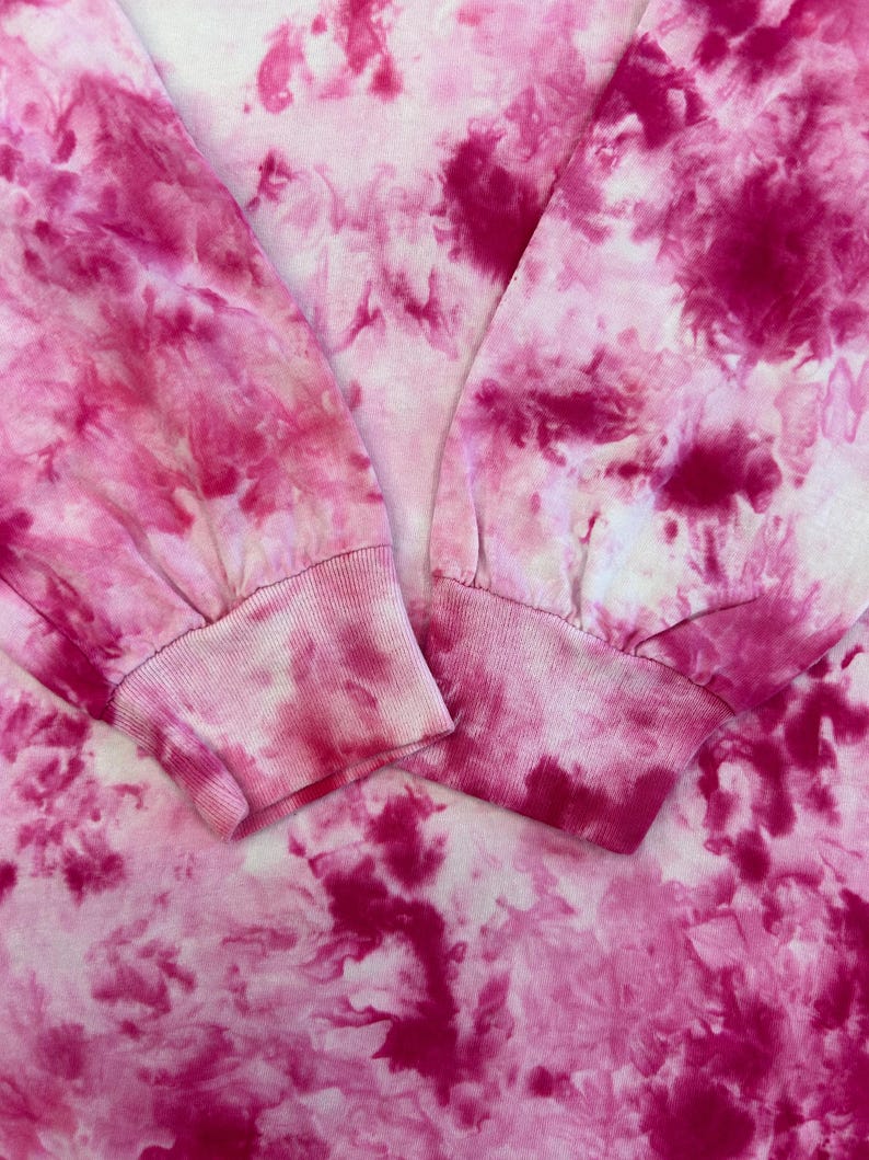 May include: Close-up of a long-sleeved shirt with a tie-dye pattern in shades of pink and white. The cuffs of the sleeves are visible, showing a ribbed texture. The fabric has a soft, blurred appearance.