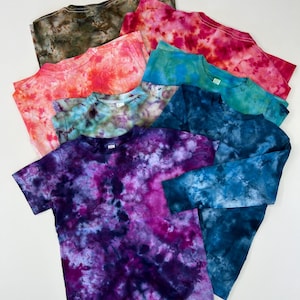 Może przedstawiać: Kolekcja koszulek typu tie-dye w różnych kolorach, w tym odcieniach różu, niebieskiego, zielonego i fioletowego. Koszulki mają okrągły dekolt i krótkie rękawy, z widoczną jedną koszulką z długim rękawem. Wzór tie-dye tworzy unikalny, kolorowy design na każdej koszulce.