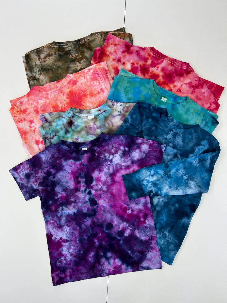 May include: A collection of tie-dye t-shirts in various colors, including shades of pink, blue, green, and purple. The shirts have a crew neck and short sleeves, with one long-sleeved shirt visible. The tie-dye pattern creates a unique, colorful design on each shirt.