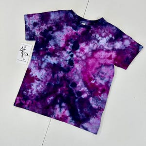 May include: A short-sleeved t-shirt with a tie-dye pattern in shades of purple, pink, and blue. The shirt has a crew neck and a tag with text on it. The tie-dye design creates a unique, colorful look.