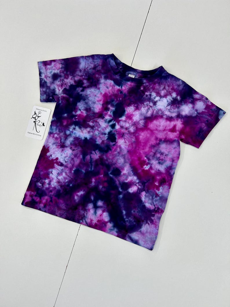 May include: A short-sleeved t-shirt with a tie-dye pattern in shades of purple, pink, and blue. The shirt has a crew neck and a tag with text on it. The tie-dye design creates a unique, colorful look.
